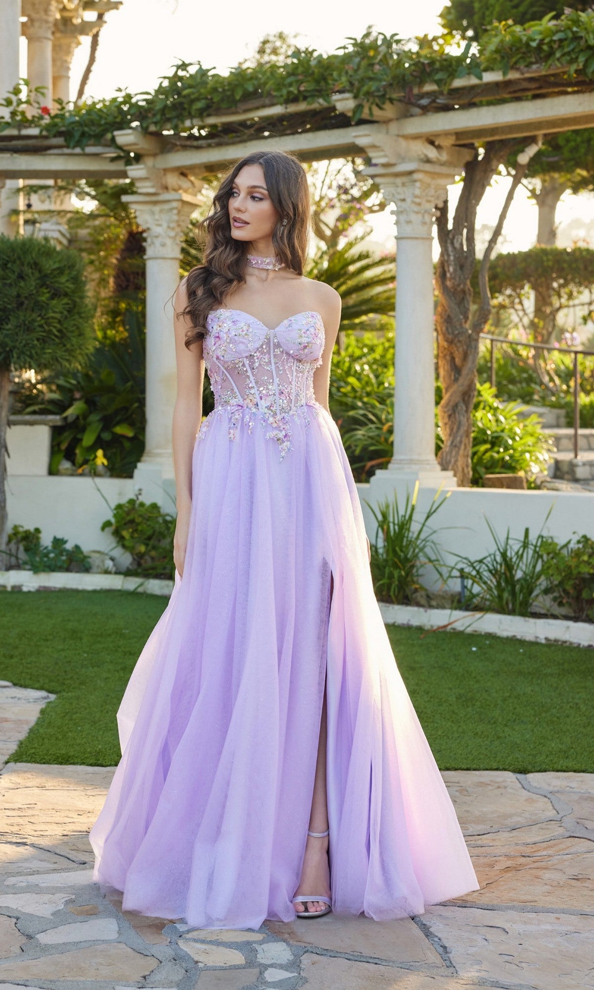 Wearing the Juliet JT2682A long lilac prom dress with a strapless glitter design and floral embroidered corset, a woman stands outside on a stone path surrounded by greenery.